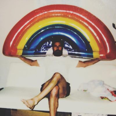 Kyan is sitting while there is a rainbow design in the back.